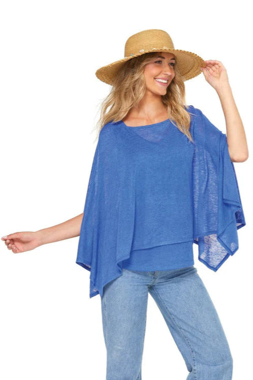 3/4 shot of a woman wearing the Centos Cami by Yul Voy in Blue, a lightweight knit tank with a v-neck and wide straps. She is wearing it  with the Cathiana Poncho and jeans and is standing in front of a white background. 