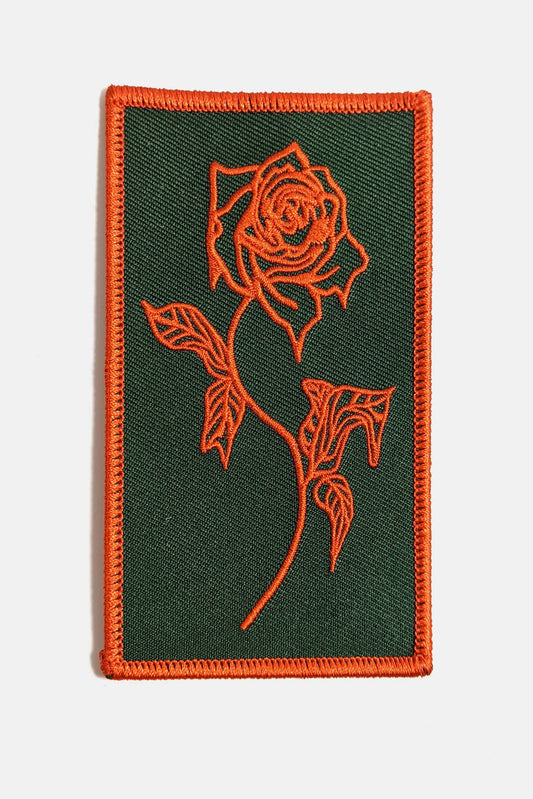 An embroidered rectangular patch from Badaboöm Studio, featuring a red Rose design on a black background, shown against a light grey background.