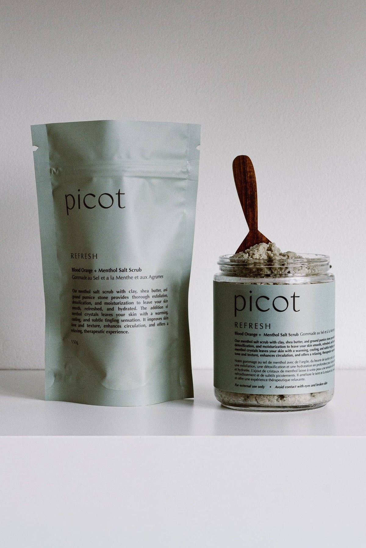 Picot Refresh salt scrub shown with its silver foil pouch and a clear glass jar containing the scrub with a wooden spoon, set against a plain white background.