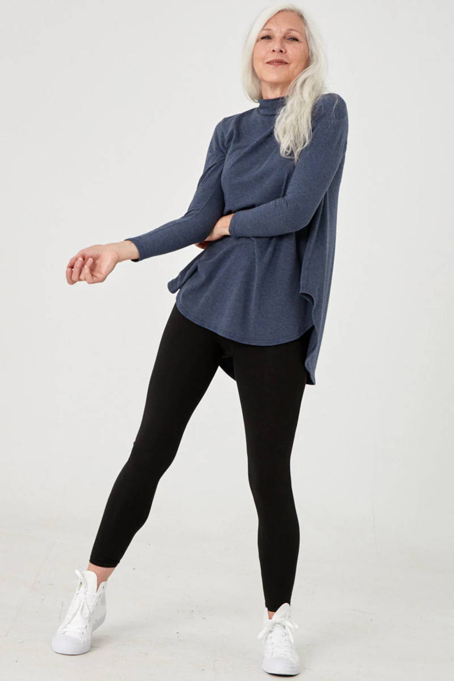 A woman wearing the Lauren Top by Advika in Blue Steel, with a turtleneck, as wing shape, long sleeves, and a rounded hi-low hem, with black leggings. 