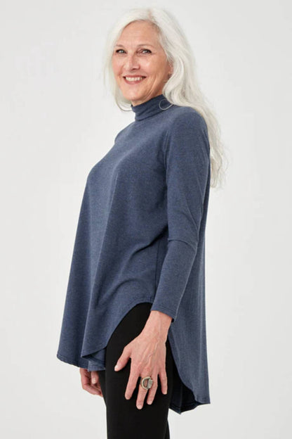 Side view of a woman wearing the Lauren Top by Advika in Blue Steel, with a turtleneck, as wing shape, long sleeves, and a rounded hi-low hem, with black leggings. 