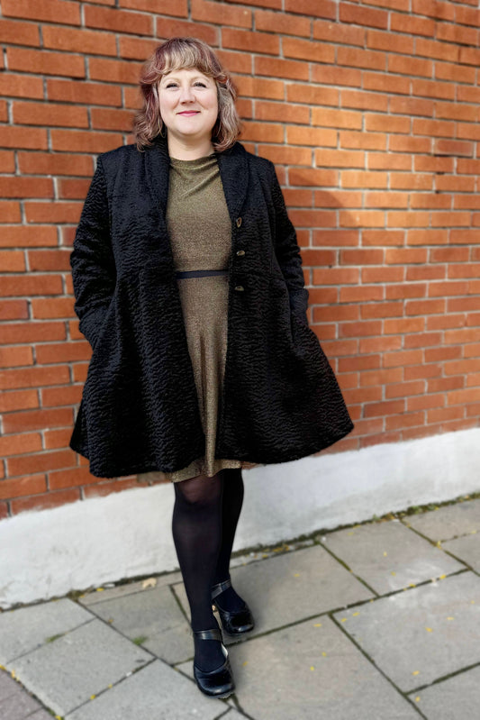 A woman wearing the Miyuki Dress by Infime in Gold, with the  Spies Jacket by Luc Fontaine, standing in front of a brick wall 