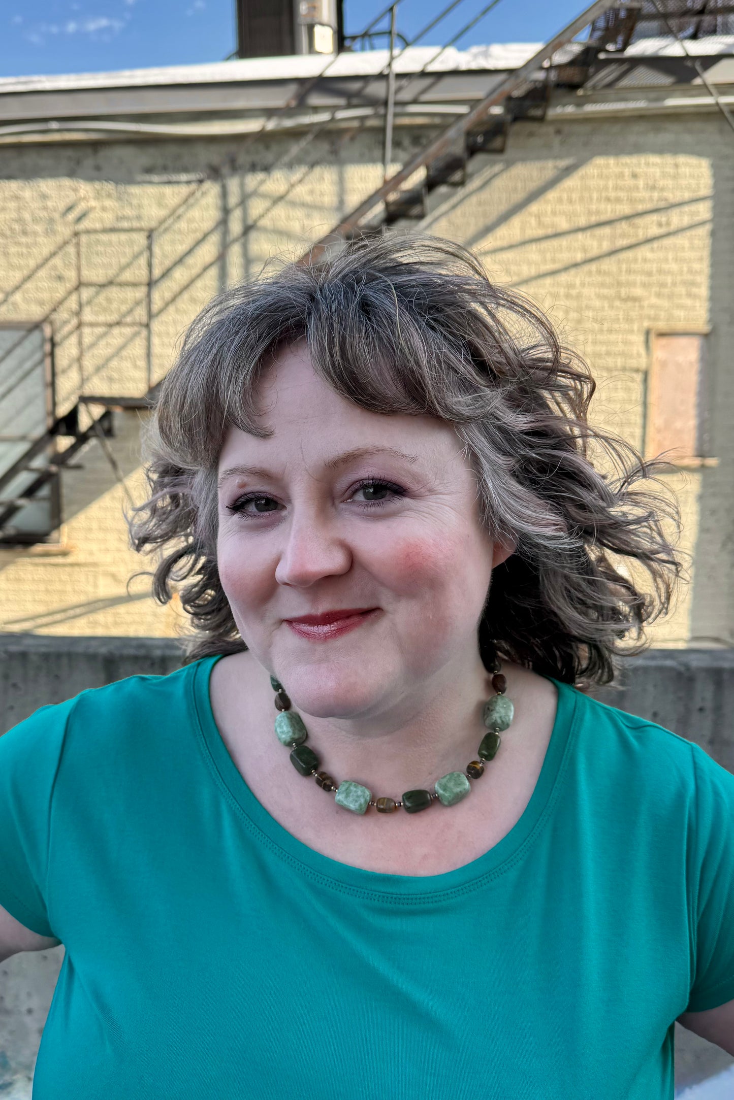 A close up of a woman wearing the Amy Short Sleeve Top by Moovment in Aqua, a t-shirt with a wide neck and short sleeves in bamboo viscose. She is standing outside by a brick wall. 