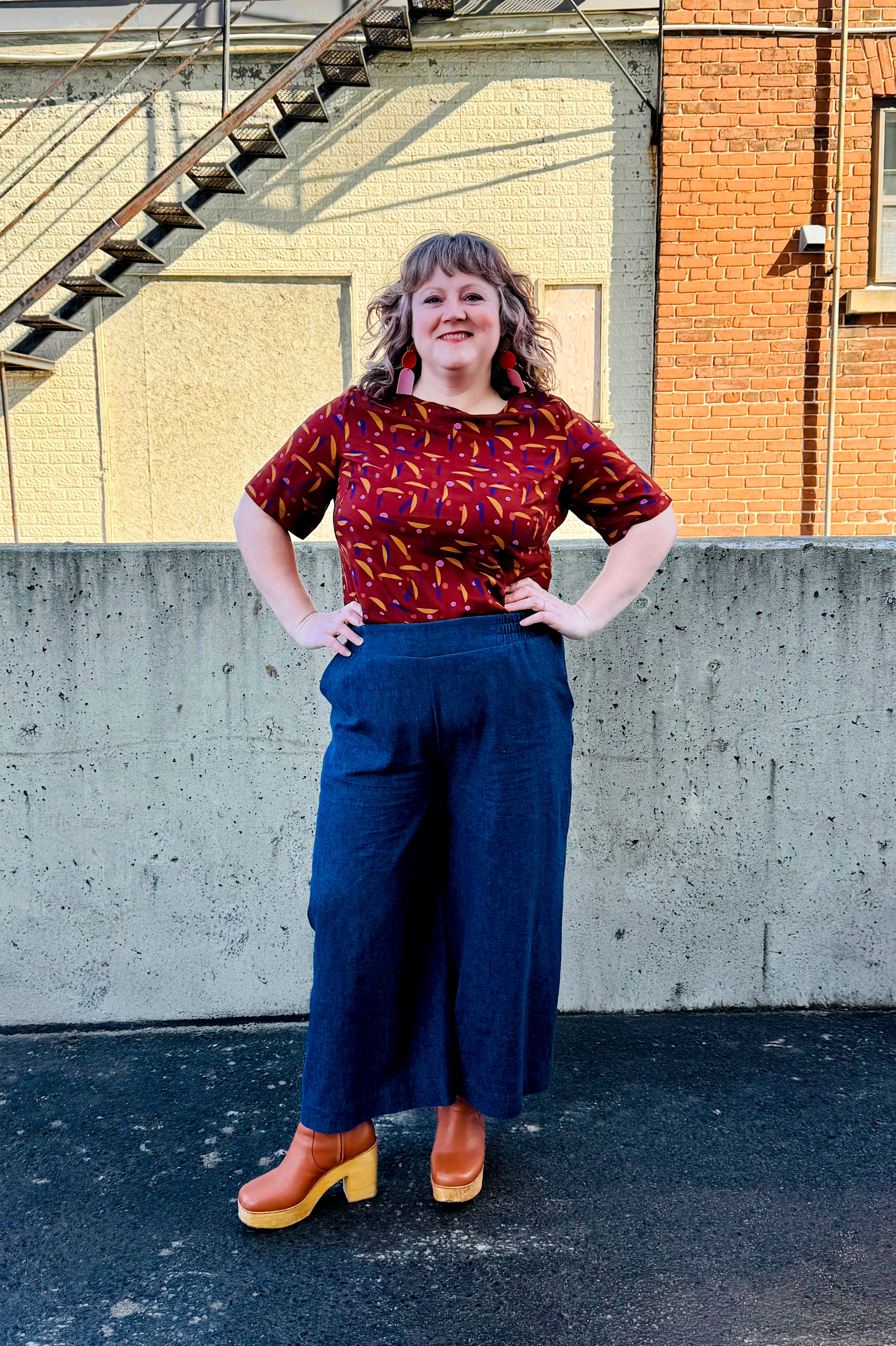 A woman wearing the Mateo Pants by Melow in Denim, ¾ wide leg pants with pull-on waistband that’s elasticized at the back. She is wearing them with the Cecil Box Blouse by Mandala and standing outside in front of a brick building. 