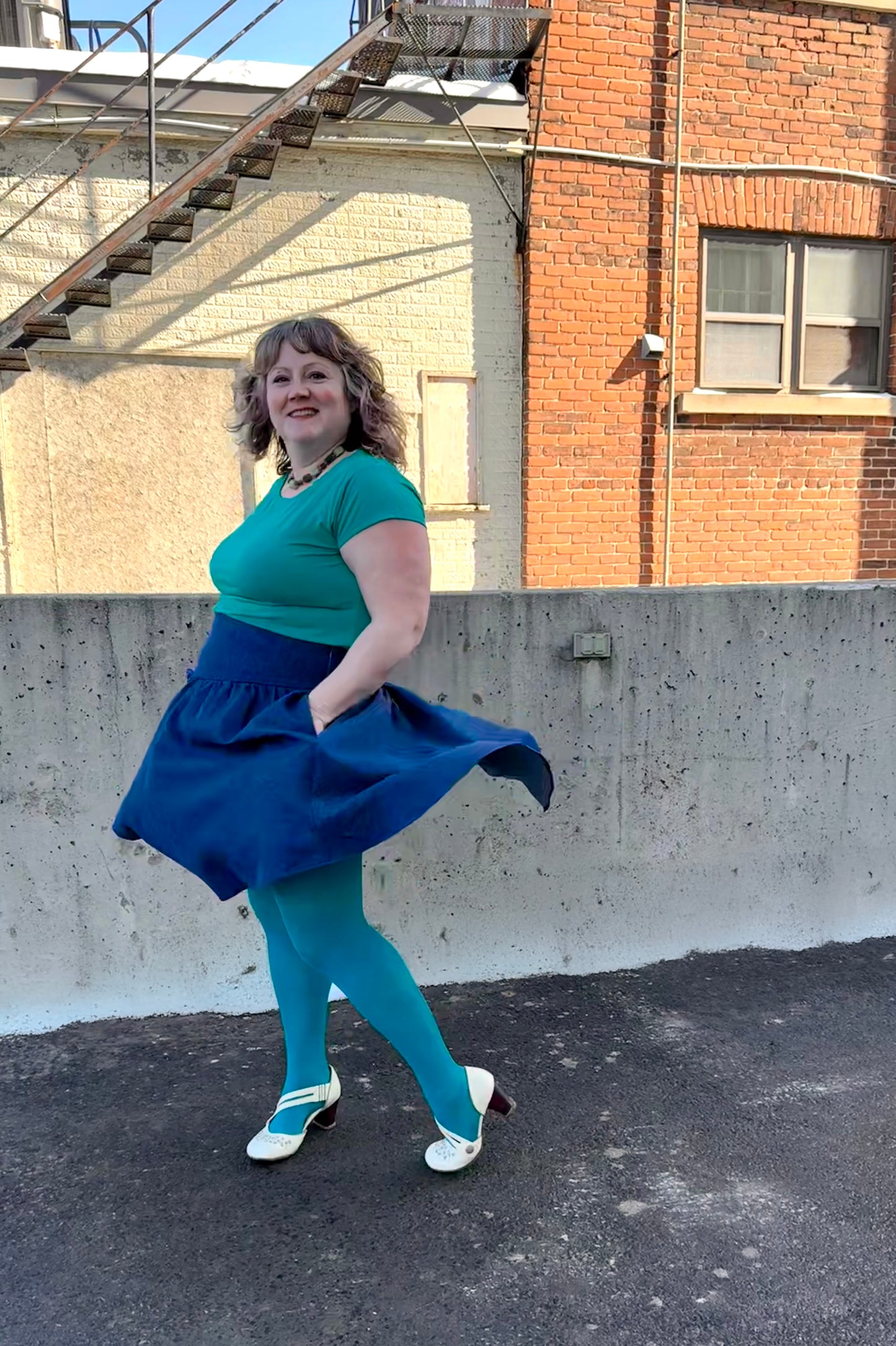 A woman wearing the Melow Margot Denim Skirt, a knee-length skirt with a wide pointed waistband, A-line shape, and invisible side zipper. She is wearing it with a turquoise top and turquoise tights and standing outside in fronnt of a brick building. 