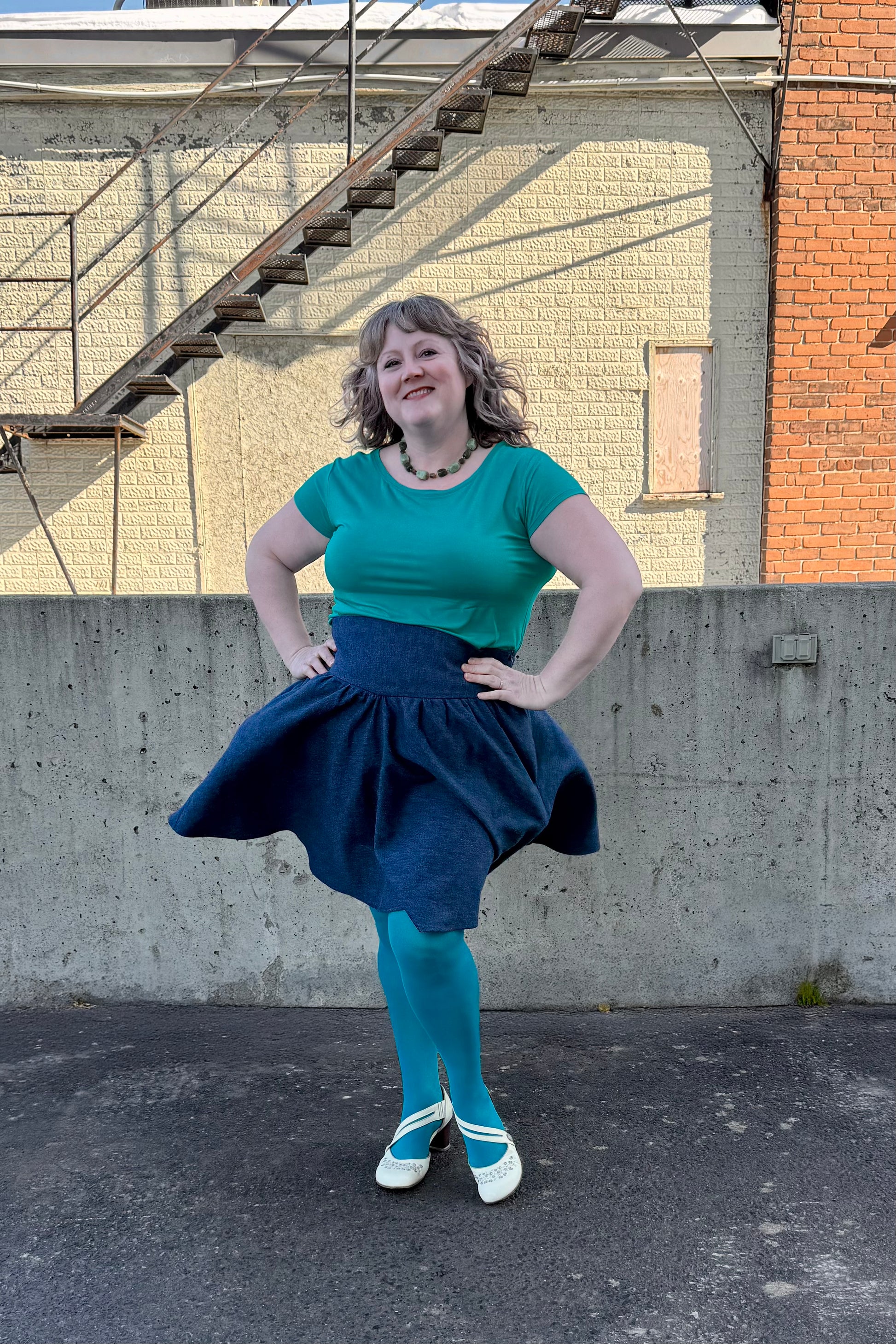 A woman wearing the Melow Margot Denim Skirt, a knee-length skirt with a wide pointed waistband, A-line shape, and invisible side zipper. She is wearing it with a turquoise top and turquoise tights and standing outside in front of a brick building. 