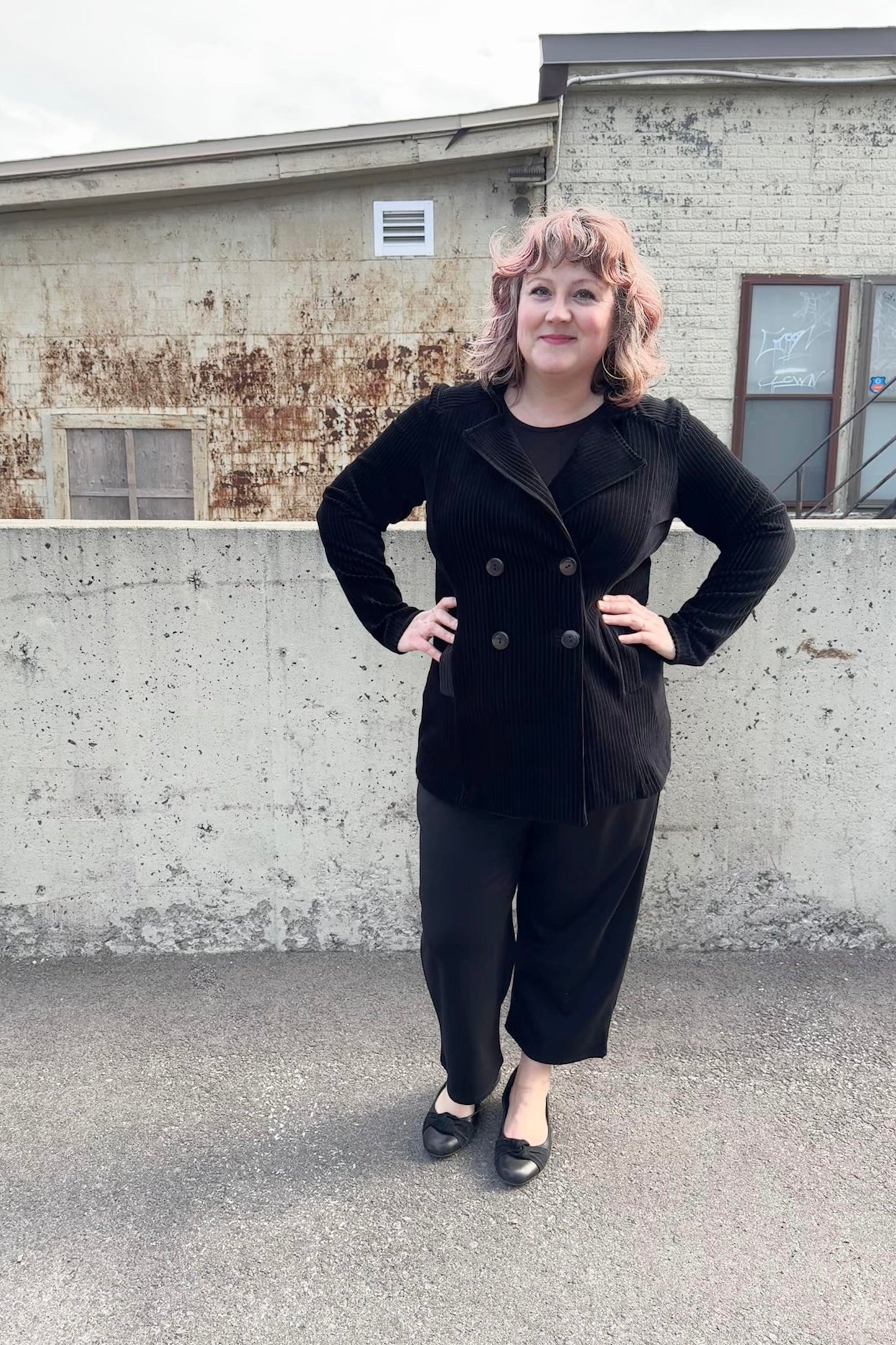 A woman wearing the Mission Blazer by Luc Fontaine in Black, in black corduroy that's slightly sheer between the wales, with a double-breasted front and front pockets. She is wearing it with black pants and standing outside in front of a brick wall. 