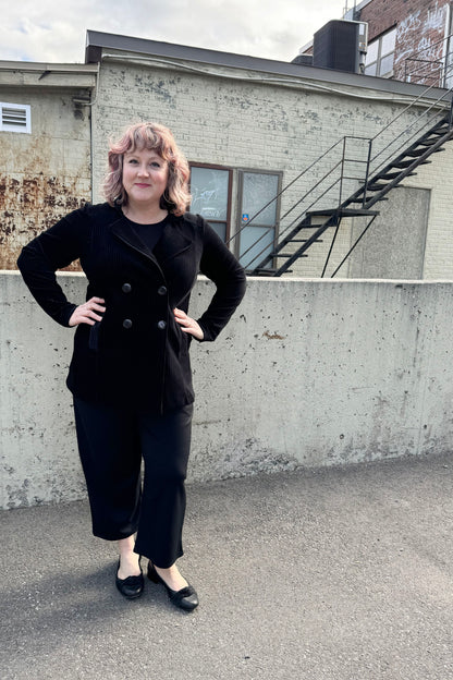 A woman wearing the Mission Blazer by Luc Fontaine in Black, in black corduroy that's slightly sheer between the wales, with a double-breasted front and front pockets. She is wearing it with black pants and standing outside in front of a brick wall. 