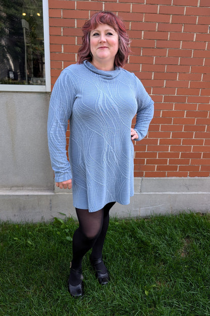 A woman wearing the Frisket Tunic by Luc Fontaine in Blue, featuring a textured fabric, cowl neck, long sleeves, and mid-thigh length, standing outside on the grass in front of a brick wall. 