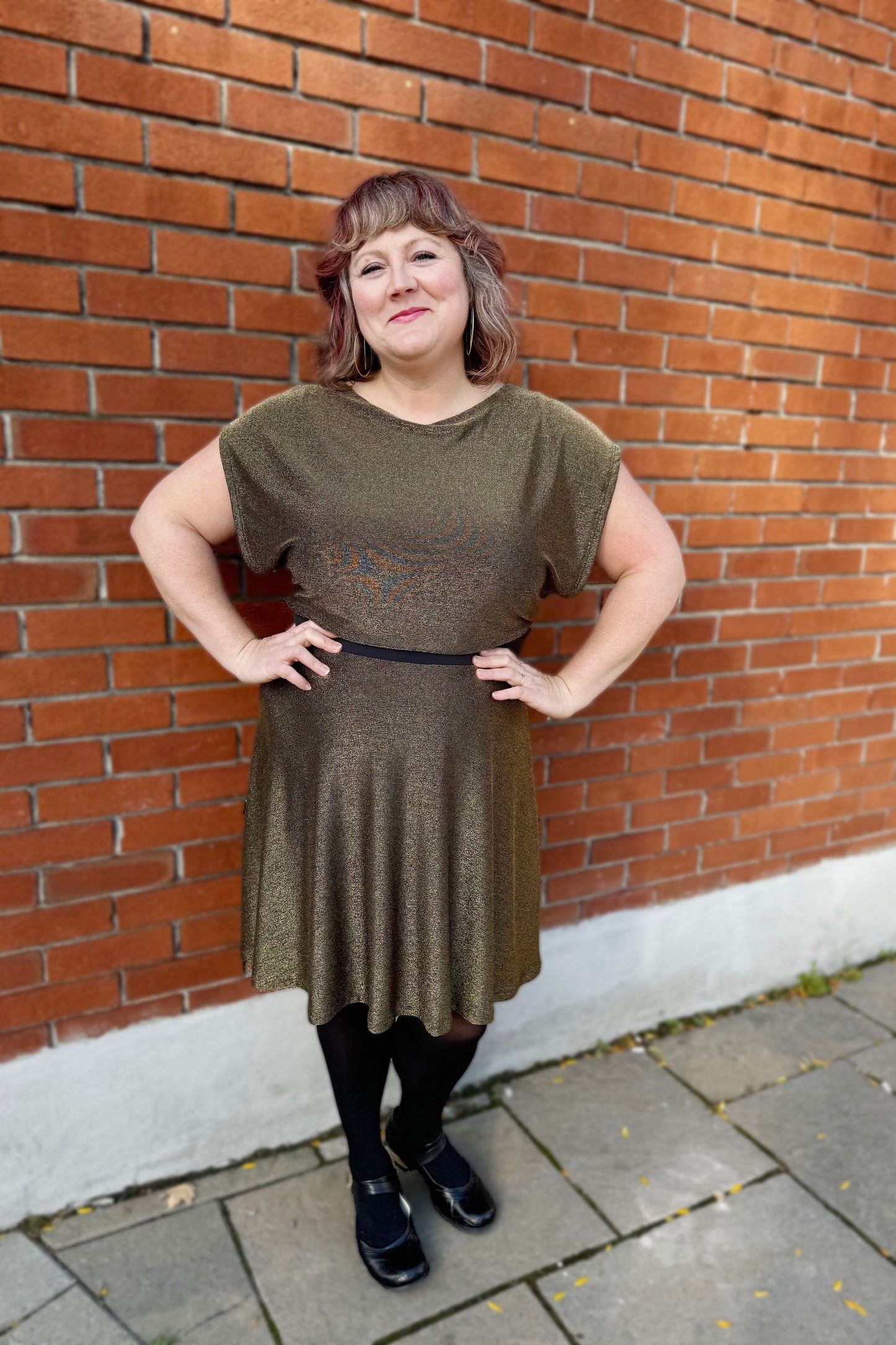 A woman wearing the Miyuki Dress by Infime in Gold, with glittery fabric, a boat neck, short extended sleeves, a contrast waistband, and a flared knee-length skirt, standing in front of a brick wall