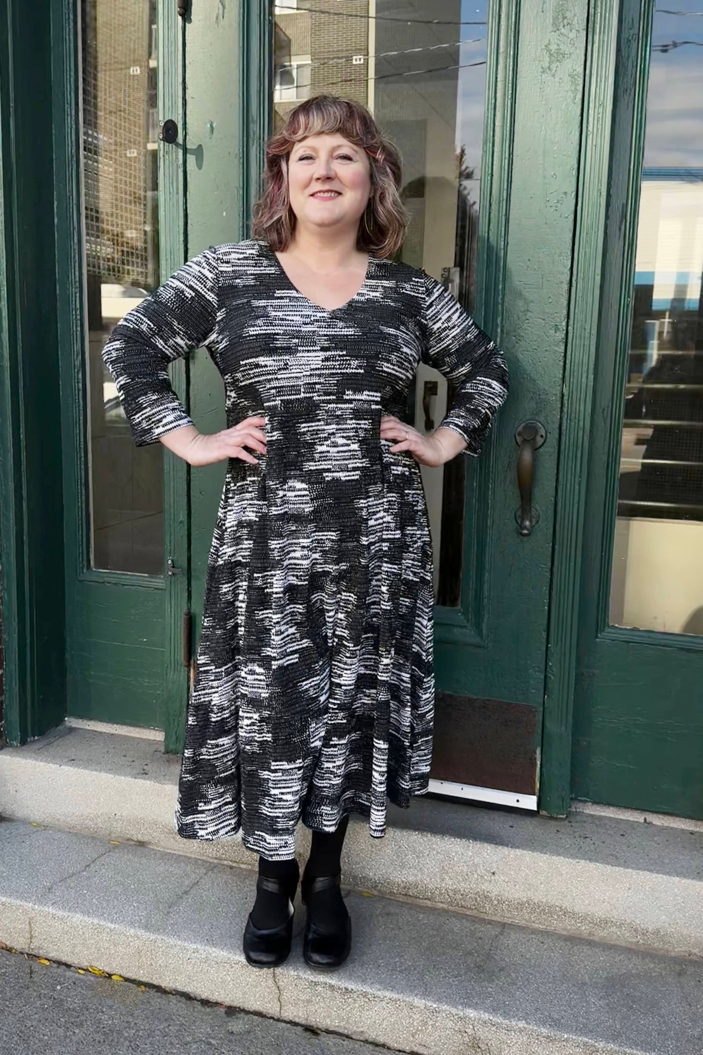 A woman wearing the Duke Dress by Pure Essence in Black/Tan, featuring a V-neck, long sleeves, a gathered waist, and a long tiered skirt, standing in front of a green doorway 