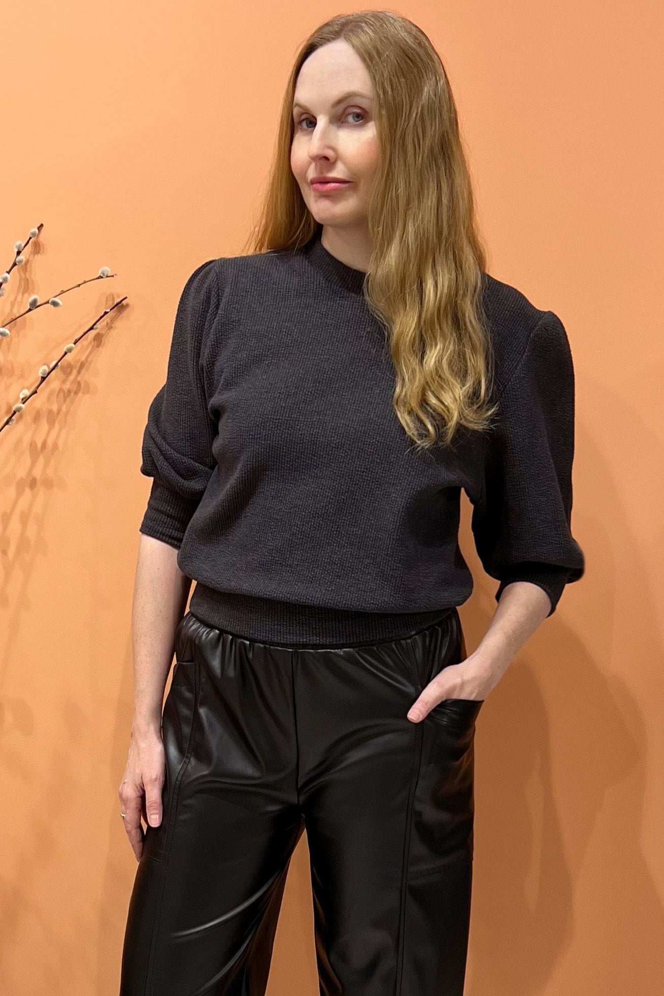 A woman wearing the Bridge Sweater by Allison Wonderland in Grey, featuring elbow length sleeves and a ribbed neckline, cuff, and hem. She's wearing it with faux leather pants and standing in front of an orange wall. 