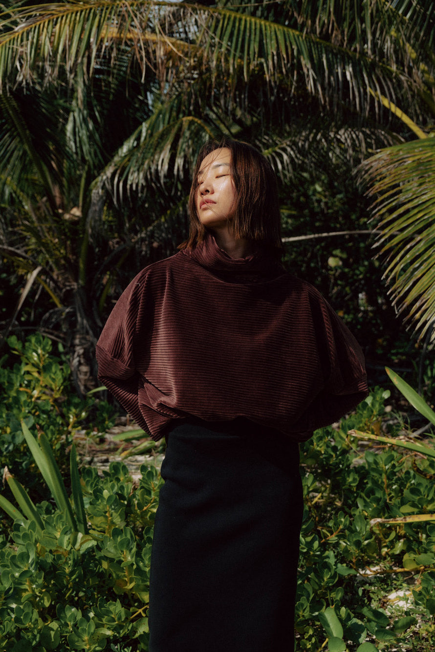 A woman wearing the Amsterdam Sweater by MAS in Cacao, an oversized ribbed velvet sweater with a chimney neckline and long batwing sleeves. She is wearing it with black pants and standing in front of a forest. 