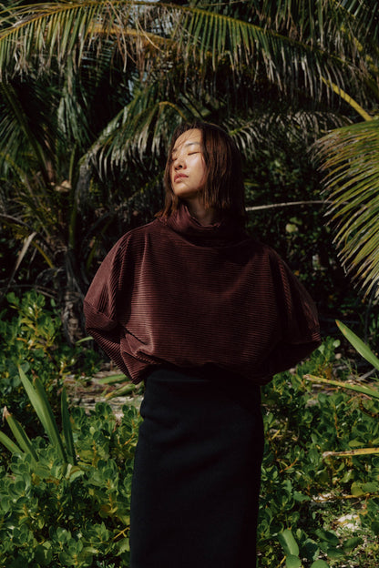A woman wearing the Amsterdam Sweater by MAS in Cacao, an oversized ribbed velvet sweater with a chimney neckline and long batwing sleeves. She is wearing it with black pants and standing in front of a forest. 