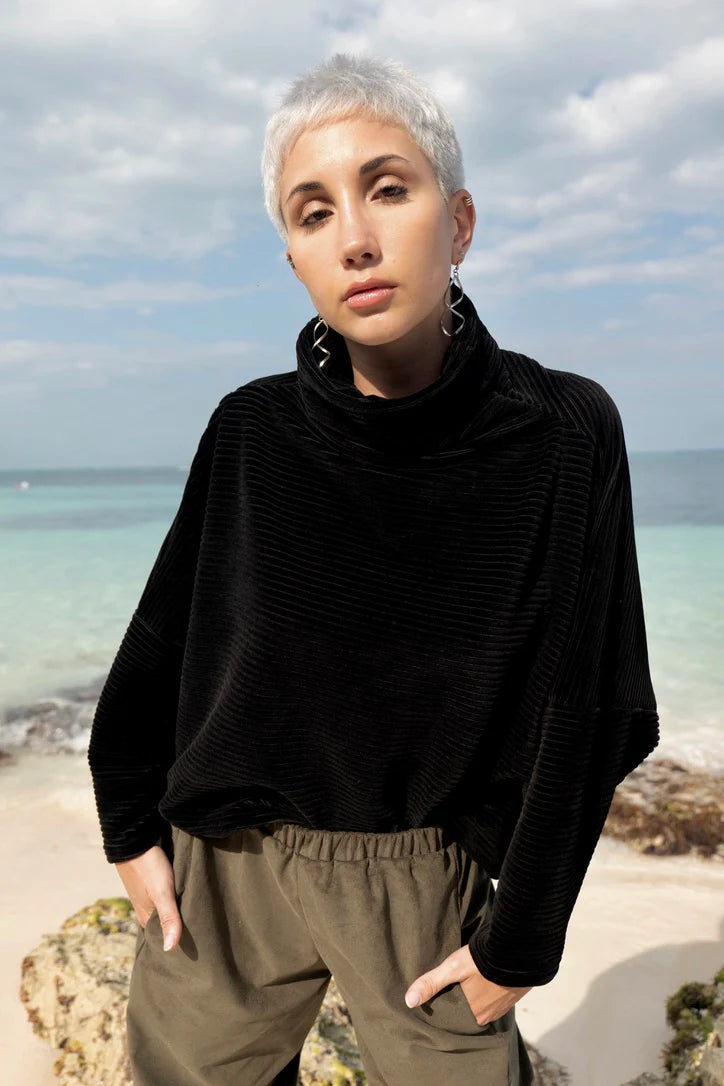 A woman wearing the Amsterdam Sweater by MAS in Pepper, an oversized ribbed velvet sweater with a chimney neckline and long batwing sleeves. She is wearing it with green pants and standing on a beach. 