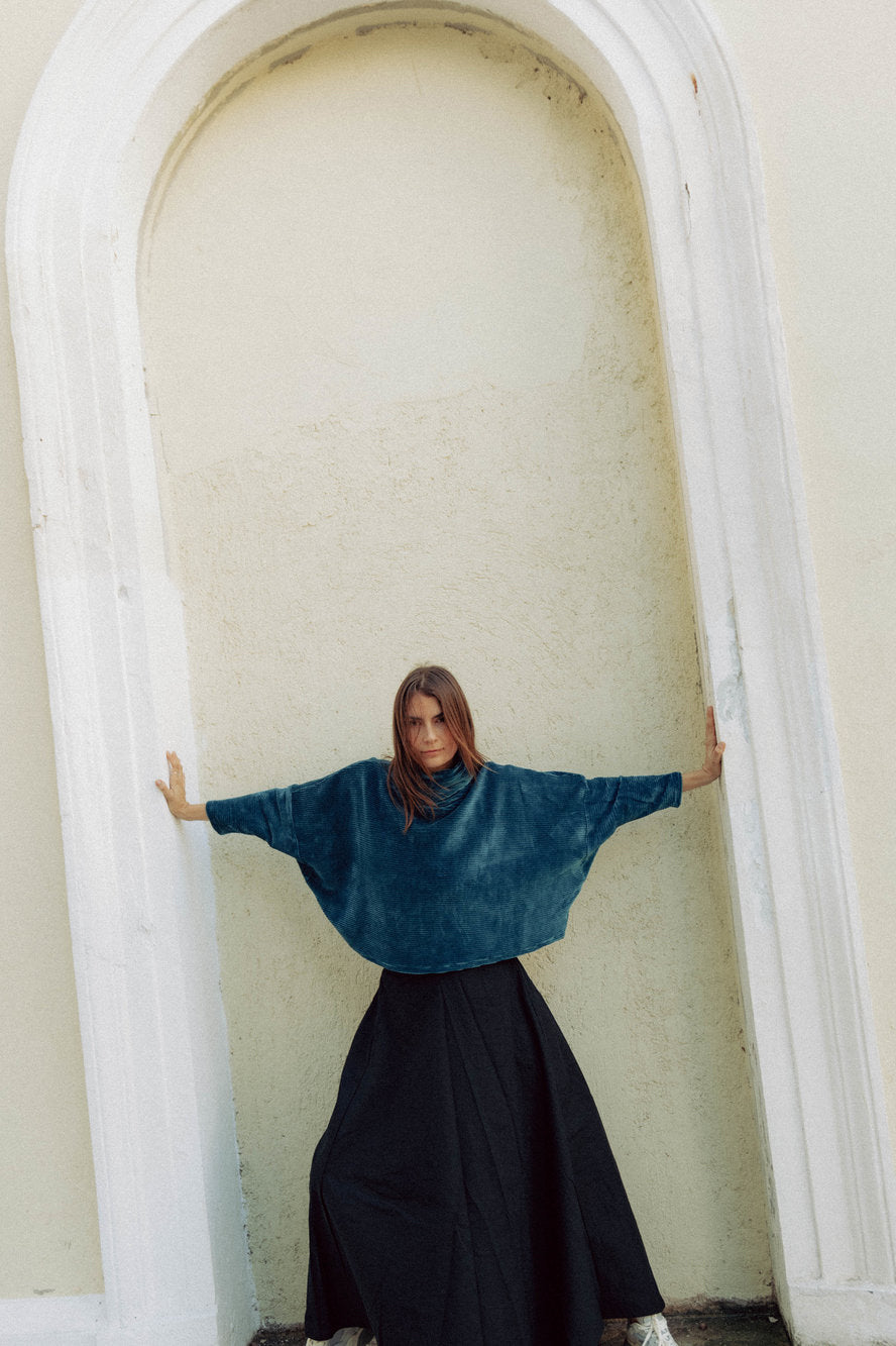 A woman wearing the Amsterdam Sweater by MAS in Juniper, an oversized ribbed velvet sweater with a chimney neckline and long batwing sleeves. She is wearing it with a black skirt and standing outside in a doorway.