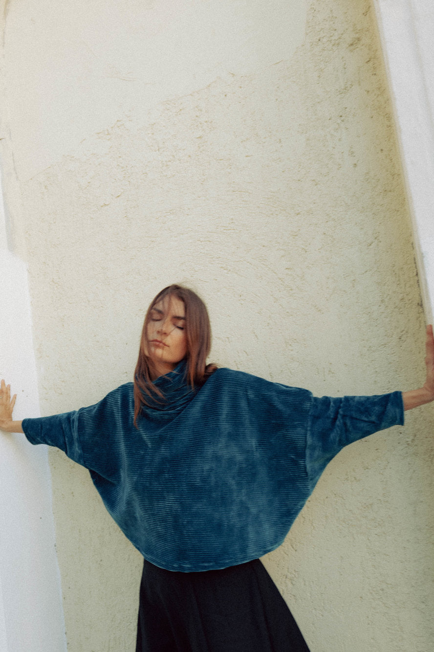 A woman wearing the Amsterdam Sweater by MAS in Juniper, an oversized ribbed velvet sweater with a chimney neckline and long batwing sleeves. She is wearing it with a black skirt pant and standing against a white wall. 