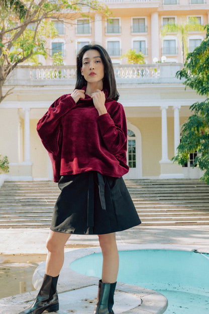 A woman wearing the Amsterdam Sweater by MAS in Rosewood, a an oversized ribbed velvet sweater with a chimney neckline and long batwing sleeves. She is wearing it with a black skirt and standing by a fountain.