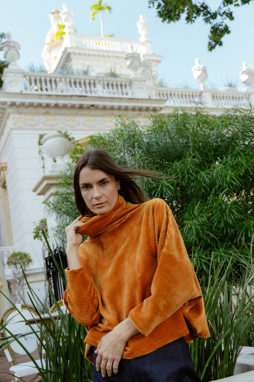 A woman wearing the Amsterdam Sweater by MAS in Tumeric, an oversized ribbed velvet sweater with a chimney neckline and long batwing sleeves. She is wearing it with blue pants and standing in a garden. 