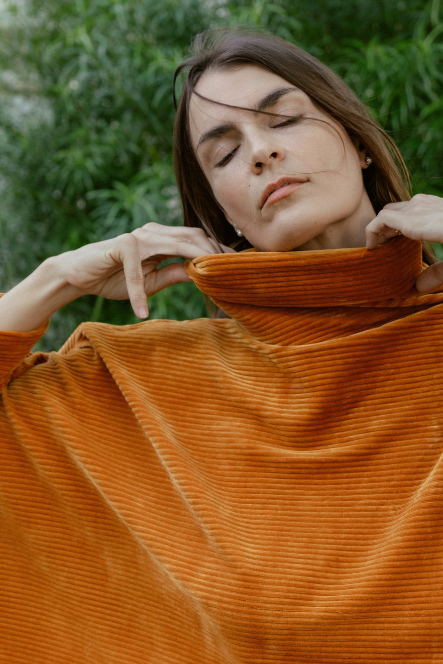 A woman wearing the Amsterdam Sweater by MAS in Tumeric, an oversized ribbed velvet sweater with a chimney neckline and long batwing sleeves.