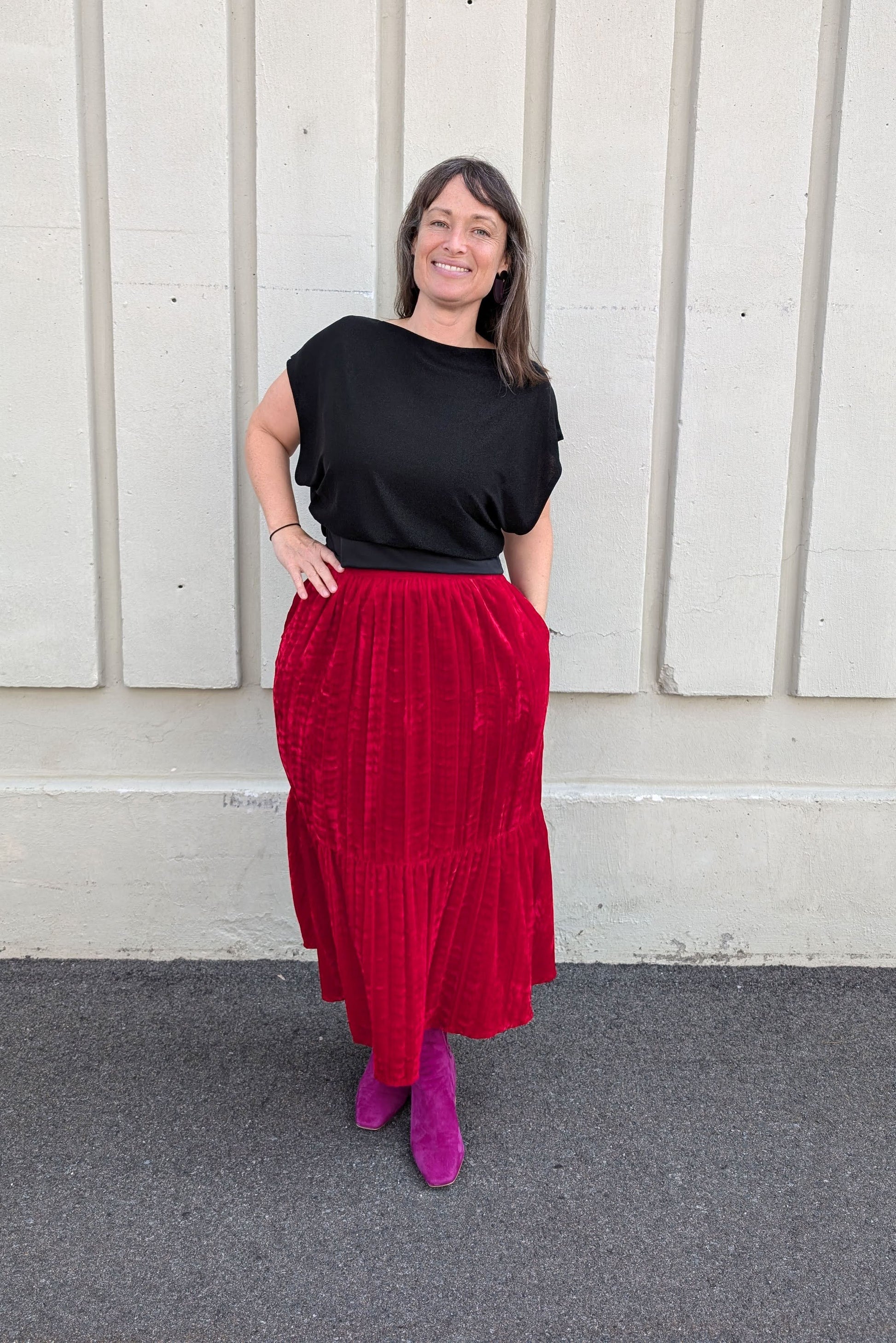 A woman wearing the Infime Saki Top in Black with the Allison Wonderland Thornbirds Skirt in Red, standing in front of a white wall 