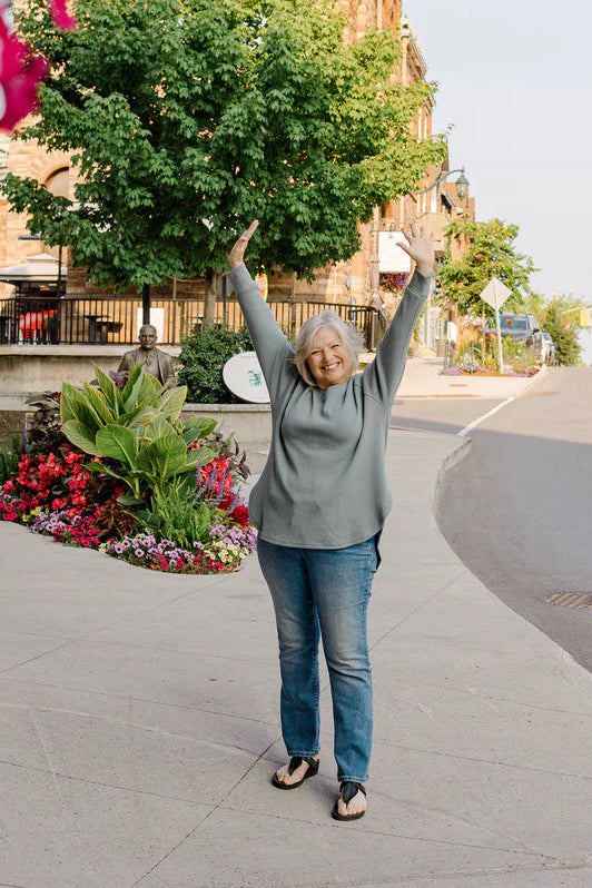 A woman wearing the Willow Waffle Sweater by Blondie in Mist, with long raglan sleeves, a loose fit, a v-neck, and side slits. She is wearing it with jeans and standing in the street with her arms raised. 