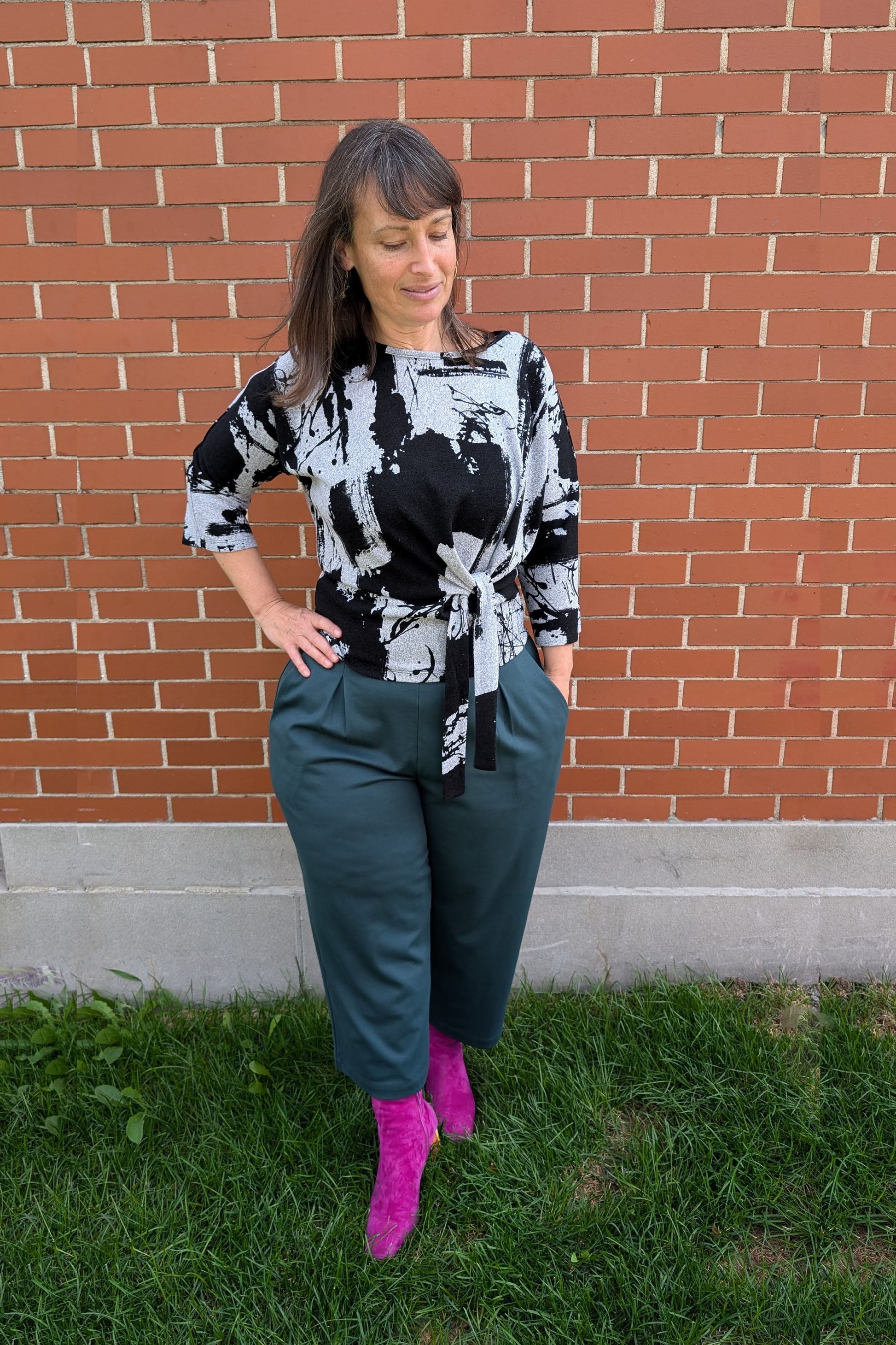 A woman wearing the NIna Top by Compli K with the Wilfred Pants by Melow in Forest - featuring 
a wide pull-on waistband, front pleats, and cropped legs, standing in front of a brick wall.