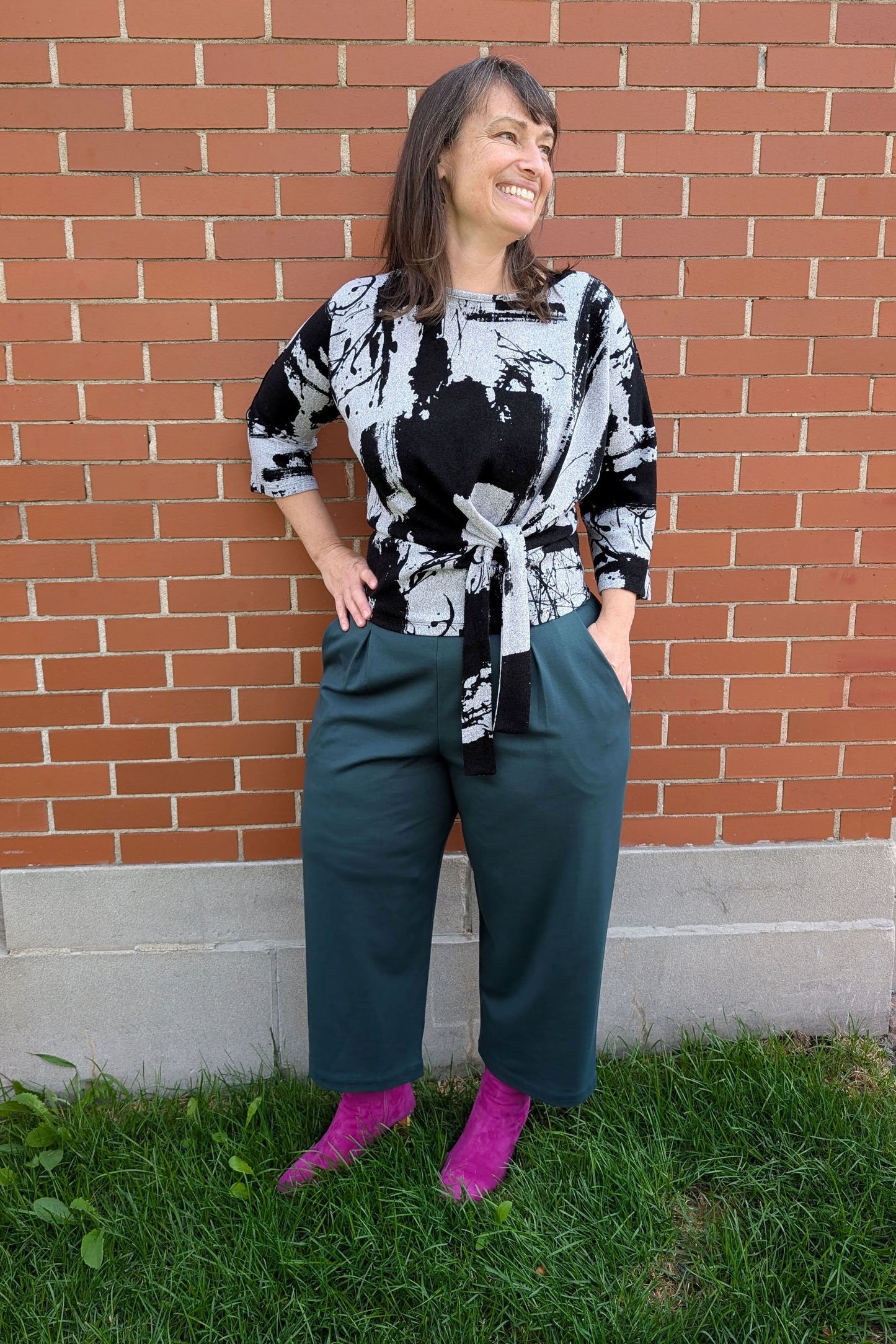 A woman wearing the NIna Top by Compli K with the Wilfred Pants by Melow in Forest - featuring 
a wide pull-on waistband, front pleats, and cropped legs, standing in front of a brick wall.