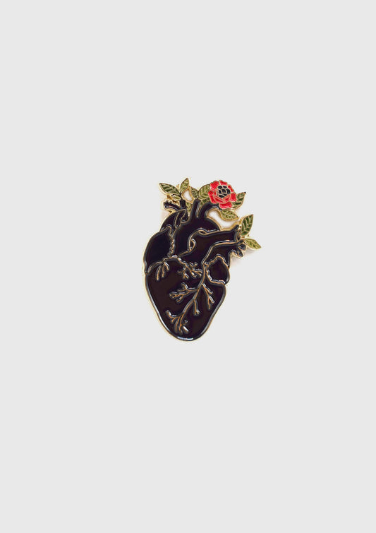 The Heart Flower Pin by Badaboöm Studio, featuring a black anatomically correct heart with flowers growing out of it, is shown against a white background. 