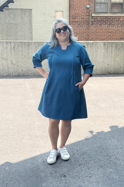 A woman wearing the Yvette Dress by Solomia in Teal, with a notched neckline, 3/4 sleeves, pleats front and back, and an above the knee hem. She is standing outside by a cement wall. 
