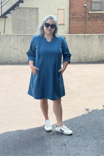 A woman wearing the Yvette Dress by Solomia in Teal, with notched neckline, 3/4 sleeves, pleats front and back, and an above the knee hem. She is standing outside by a cement wall. 