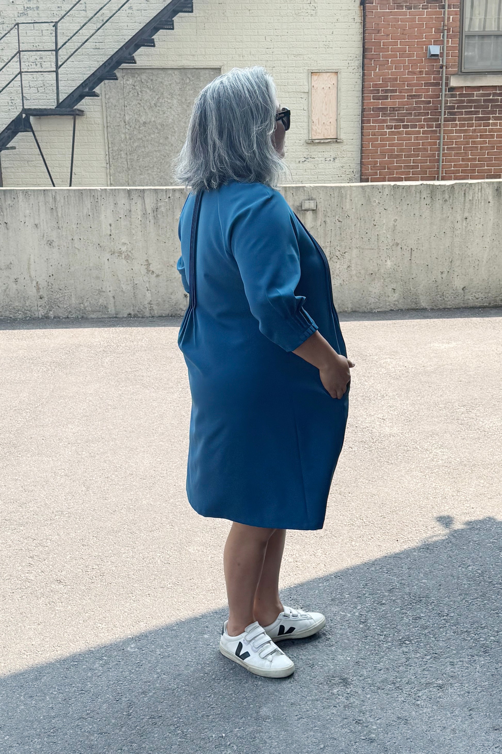 Side view of a woman wearing the Yvette Dress by Solomia in Teal, with notched neckline, 3/4 sleeves, pleats front and back, and an above the knee hem. She is standing outside by a cement wall. 