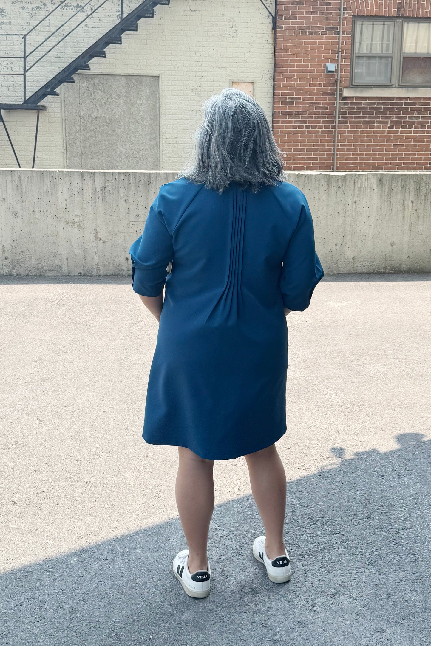 Back view of a woman wearing the Yvette Dress by Solomia in Teal, with notched neckline, 3/4 sleeves, pleats front and back, and an above the knee hem. She is standing outside by a cement wall. 