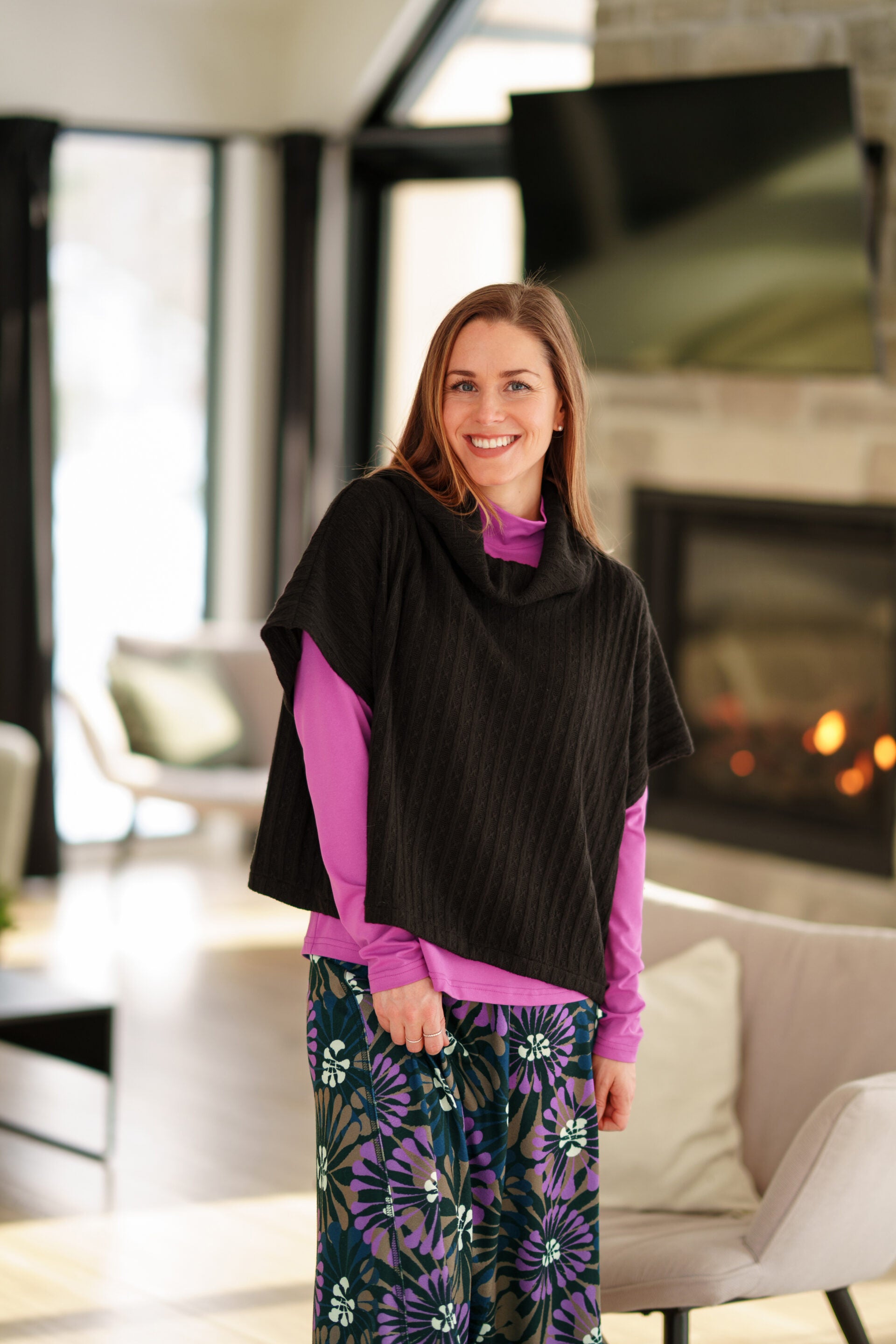 A woman wearing the Grandmont Bolero by Rien Ne Se Perd in Black,  a ribbed cowl-neck poncho. She is wearing it  over the Ste Croix Sweater in Magenta and the floral Hudson Skirt and standing in a softly lit living room with a fireplace in the background.