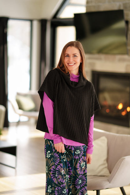 A woman wearing the Grandmont Bolero by Rien Ne Se Perd in Black,  a ribbed cowl-neck poncho. She is wearing it  over the Ste Croix Sweater in Magenta and the floral Hudson Skirt and standing in a softly lit living room with a fireplace in the background.