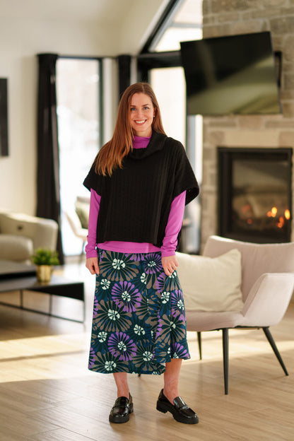 A woman wearing the Grandmont Bolero by Rien Ne Se Perd in Black,  a ribbed cowl-neck poncho. She is wearing it  over the Ste Croix Sweater in Magenta and the floral Hudson Skirt and standing in a softly lit living room with a fireplace in the background.