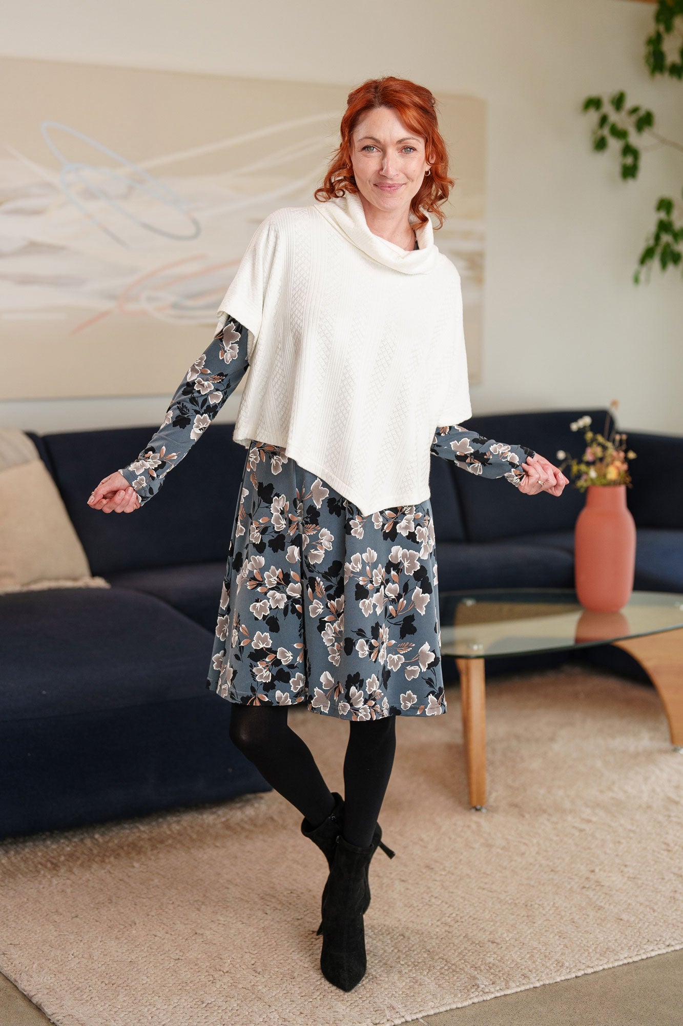 A woman wearing the Mariana Dress by Rien ne se Perd in Blue Floral, featuring a V-neck, an empire waist with a ribbon detail, long sleeves, and a knee-length A-line skirt. She is wearing it with the Lupin Bolero-Poncho in Ivory and standing in a living room setting.