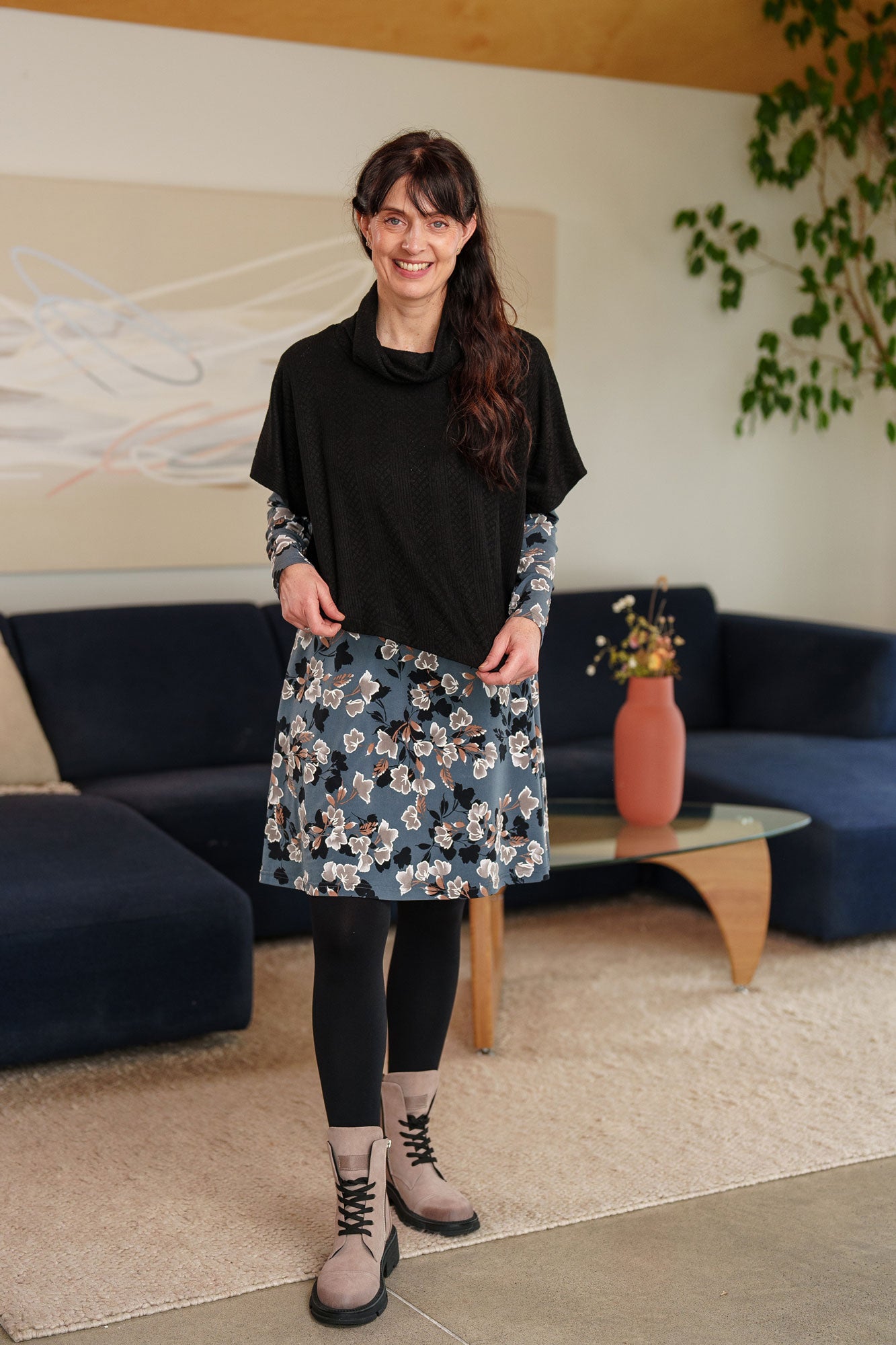 A woman wearing the Mariana Dress by Rien ne se Perd in Blue Floral, featuring a V-neck, an empire waist with a ribbon detail, long sleeves, and a knee-length A-line skirt. She is wearing it with the Lupin Bolero-Poncho in Black and standing in a living room setting.