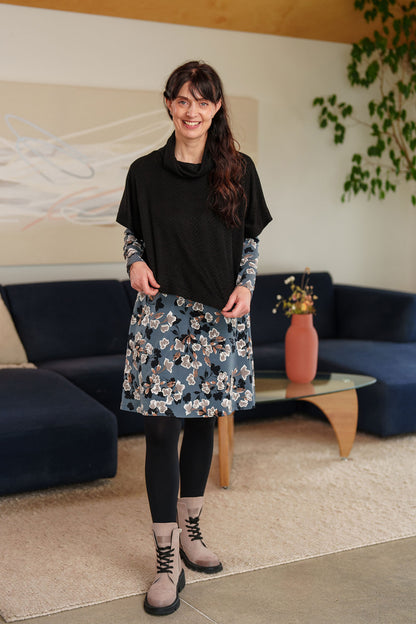 A woman wearing the Mariana Dress by Rien ne se Perd in Blue Floral, featuring a V-neck, an empire waist with a ribbon detail, long sleeves, and a knee-length A-line skirt. She is wearing it with the Lupin Bolero-Poncho in Black and standing in a living room setting.