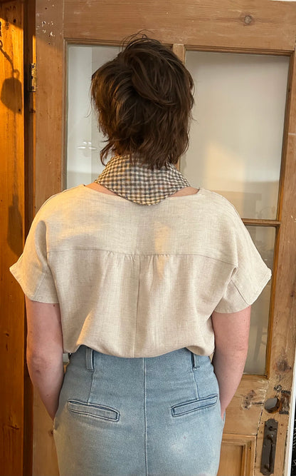 Back view of a woman wearing the Boronia Top by Kazak in Natural Linen with a round neck, short sleeves, slightly cropped and boxy fit, with a box pleat at the back yoke. She is wearing it with a checkered scarf and light jeans and standing in front of a wooden door. 