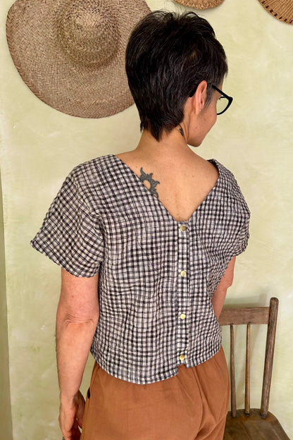 Back view of a woman wearing the Brendola Top from Kazak in Black and Cream Vichy/Gingham, a slightly cropped short sleeved top with three brass snaps that can be worn at the back or the front. She is wearing it with the Oxalis Pants in Ginger and standing in a room with a wooden chair. 