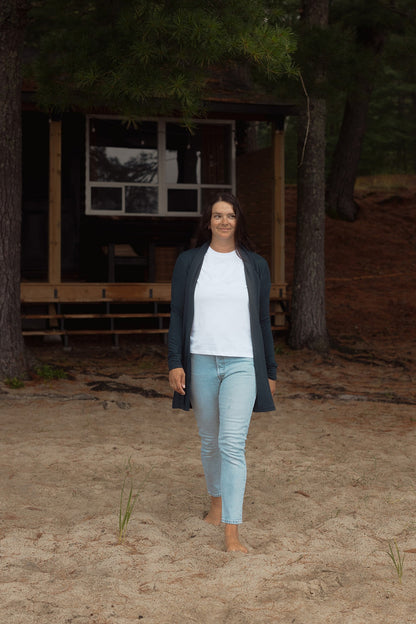 A woman wearing the Briton Cardigan from Blondie in Poppy, a mid-thigh length open cardigan with a small collar and in-seam pockets. She is wearing it with a white tee shirt and jeans and outside a cabin. 