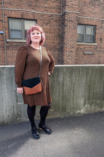 A woman wearing the Gretta Dress by Luc Fontaine in Caramel and Black print, featuring a round neck, slightly puffed sleeves, textured fabric, and above the knee length skirt, standing outside in front of a brick wall. 