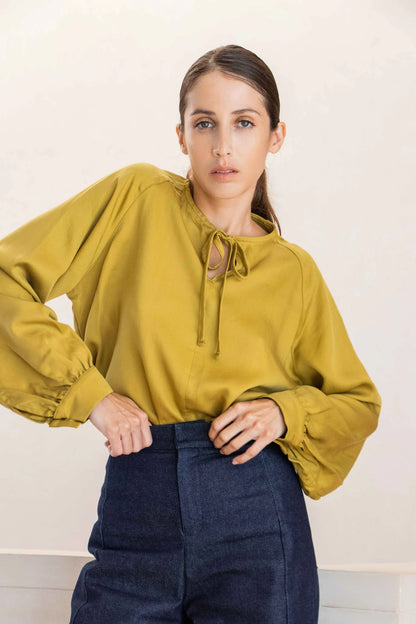 A woman wearing the Cairo Blouse by MAS in Lime, made from lyocell with raglan sleeves with gathered cuffs, and a keyhole and cord neckline. She is wearing it with the Vienna Trousers in Jeans and standing in front of a white background. 