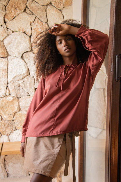 A woman wearing the Cairo Blouse by MAS in Paprika, made from lyocell with raglan sleeves with gathered cuffs, and a keyhole and cord neckline. She is wearing it with a beige skirt and standing outside by a stone wall. 