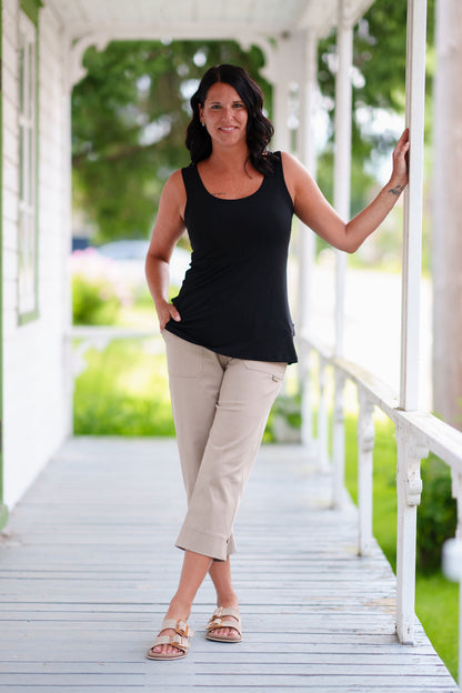 A woman wearing the Vino 3/4 Pants from Rien ne se Perd in Natural, elastic waisted pants with large pockets, and slim 3/4 legs with side slits with coconut button details. She is wearing them with a black tank and standing on a porch. 