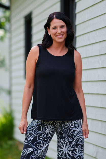 A woman wearing the Spritz Tank Top by Rien ne se Perd in Black, with wide straps and side slits. She is wearing it with  a black tank top and patterned pants and standing in front of a house.