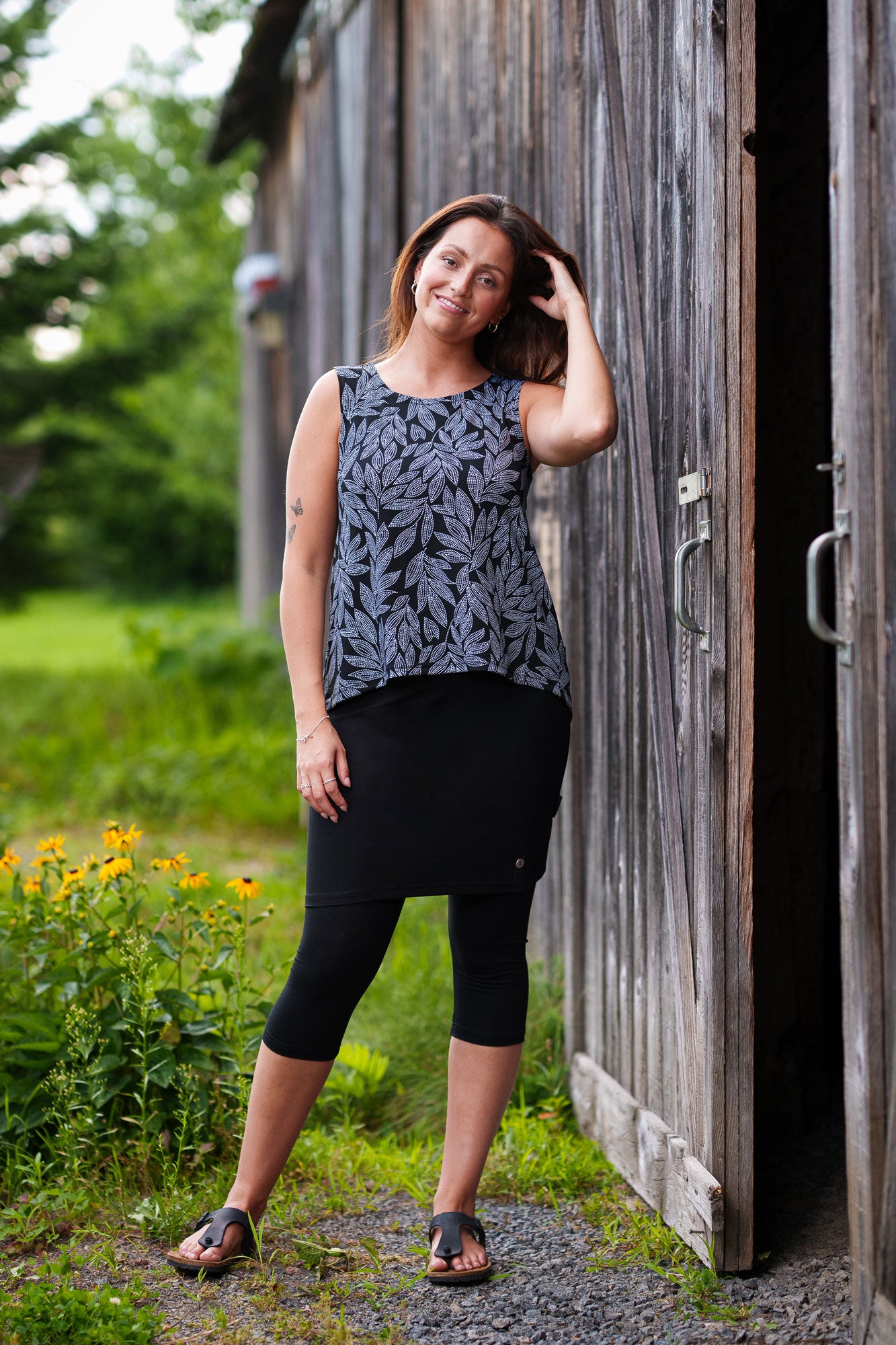 A woman wearing the Swing Cami by Rien ne se Perd in Black and White Foliage, a trapeze cut tank with a hi-low rounded hemline. She is wearing it with a black skirt and standing outside in front of a wooden building. 