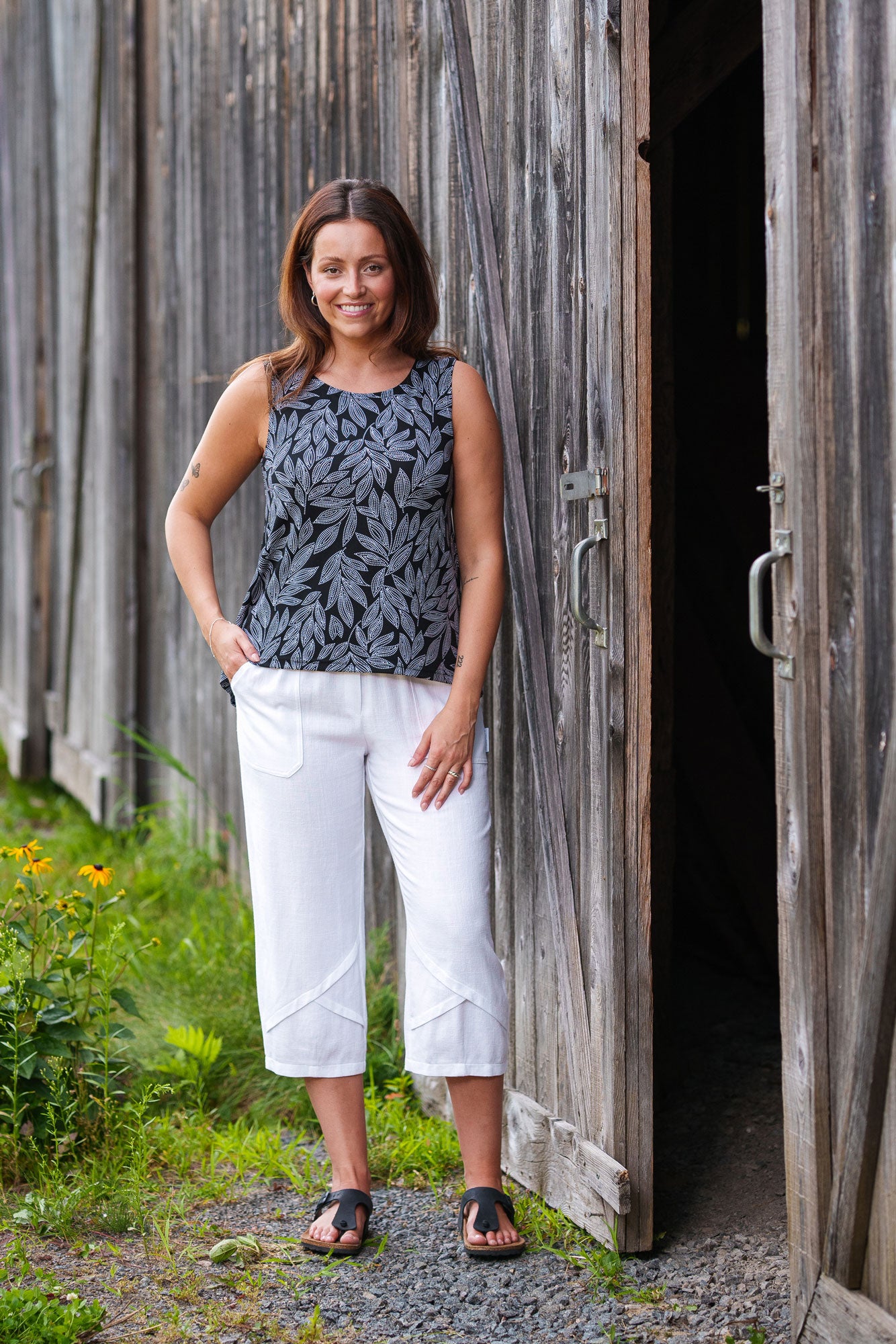 A woman wearing the Swing Cami in Black and White Foliage by Rien ne se Perd with white capri pants, standing outside in front of a wooden building. 