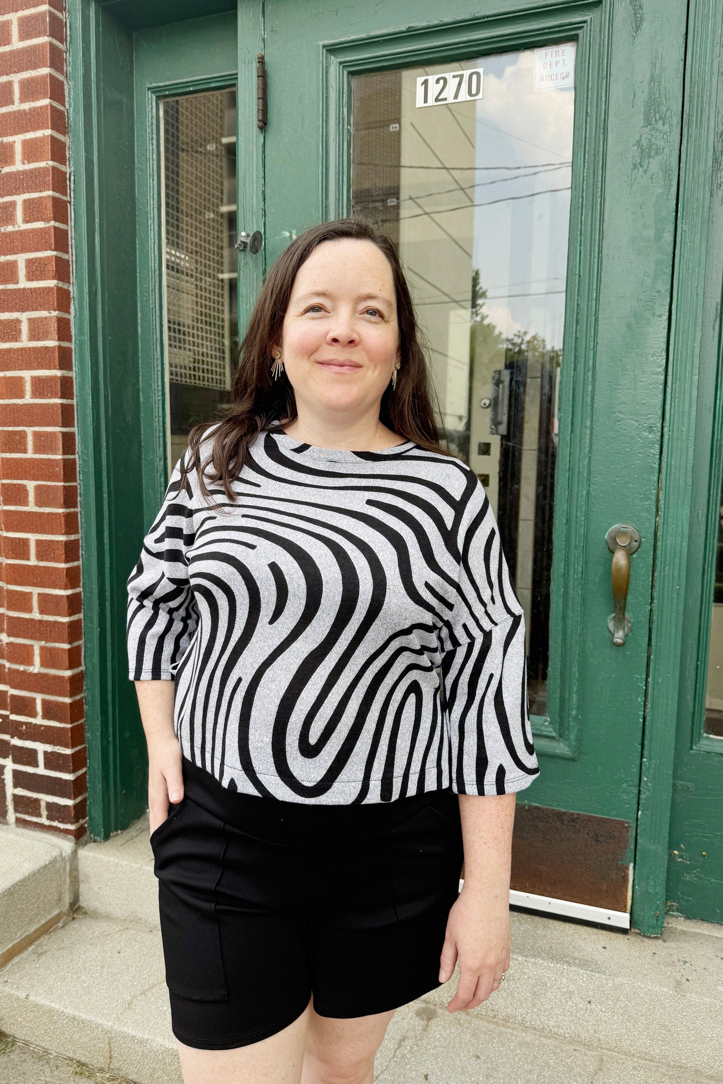 A woman wearing the Paula Top by Compli K in Silver Black Swirl, with round neck, 3/4 drop sleeves, and a hip length, with black shorts. She is standing outside in front of a door. 